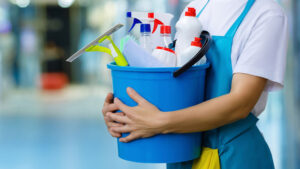 A close-up of a person wearing a white shirt and blue apron holding a blue bucket containing various cleaning supplies.